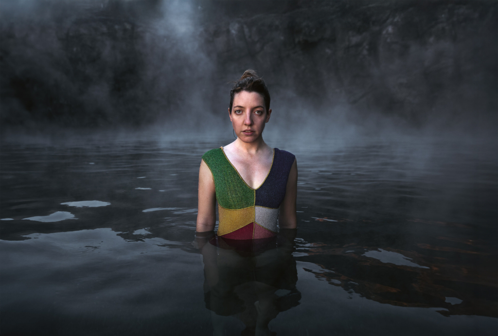 A woman stands waist-deep in dark water, facing the camera against a backdrop of mist and rocky cliffs. Wearing a colorful geometric-patterned dress, she holds a steady, thoughtful gaze as soft fog drifts across the surface, creating a quiet and atmospheric scene.