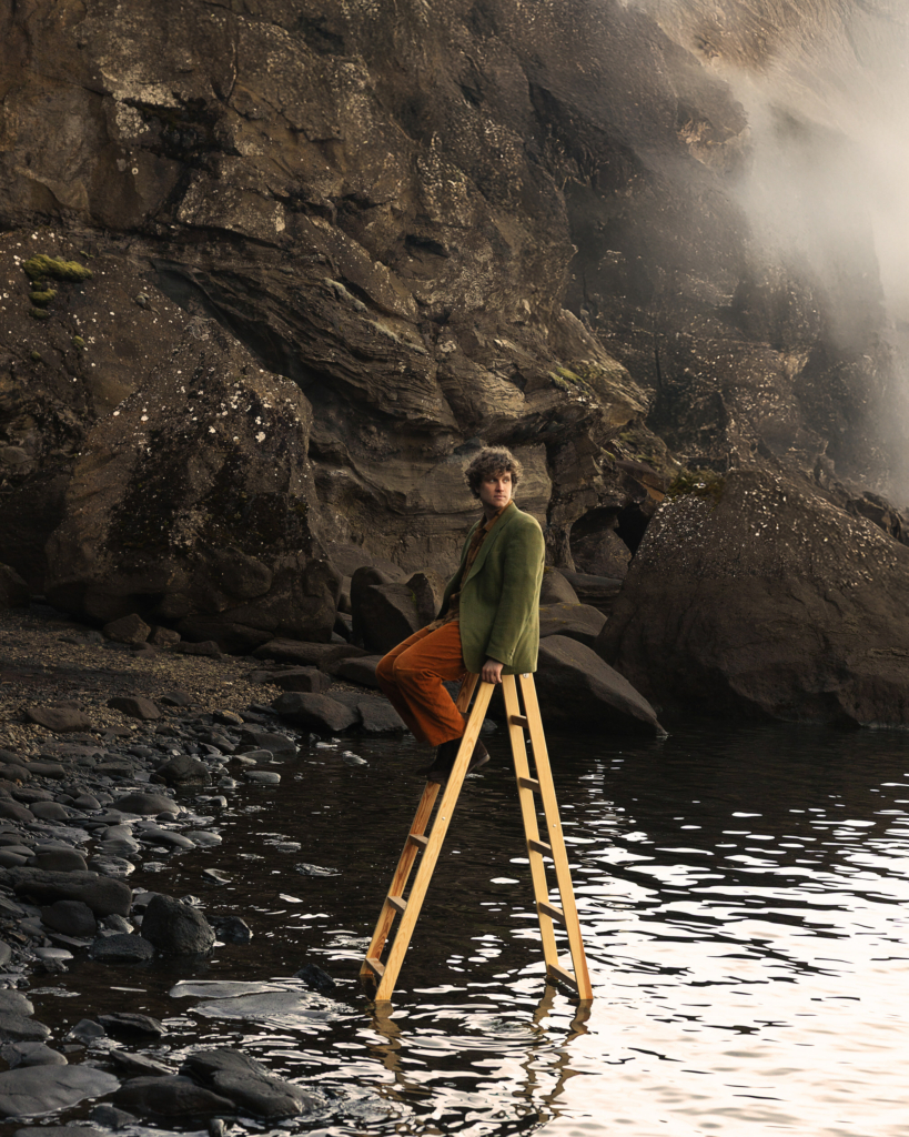 A man sits atop a wooden ladder standing in shallow water beside a rocky shoreline and towering cliff face. Dressed in earthy green and rust-colored clothing, he looks off into the distance with a thoughtful expression as mist drifts across the rugged landscape behind him.