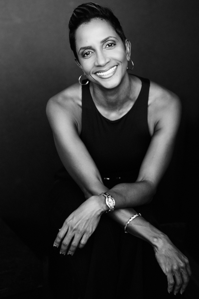 Black and white portrait of a smiling woman with short hair seated with her arms folded in front of a dark background.