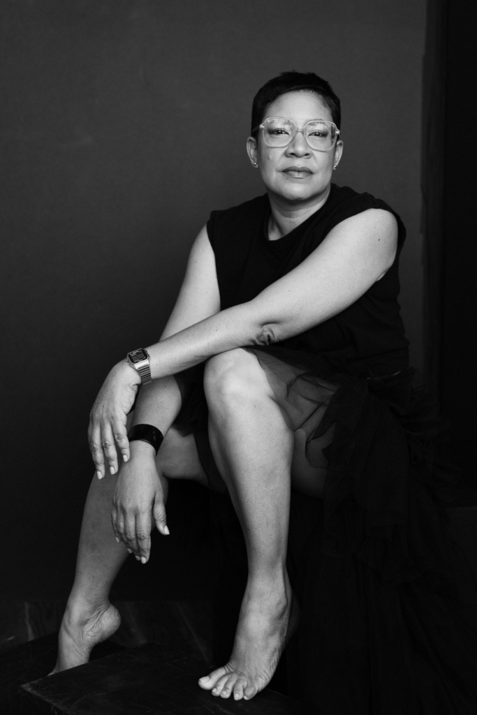 Black and white studio portrait of a barefoot woman in glasses seated in a black dress against a dark backdrop.
