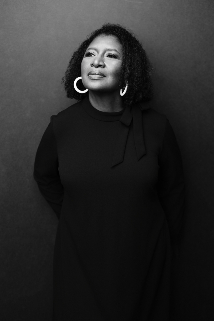 Black and white portrait of a woman in oversized white earrings gazing upward against a dark studio backdrop.