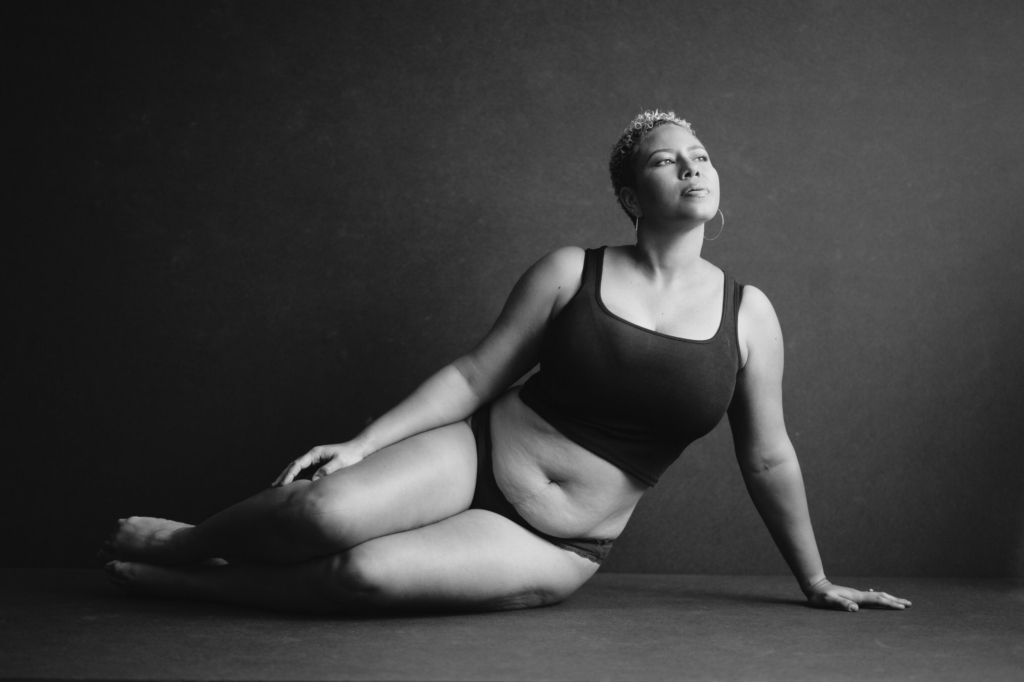 Black and white studio portrait of a woman reclining confidently against a dark backdrop during a body-positive portrait session.