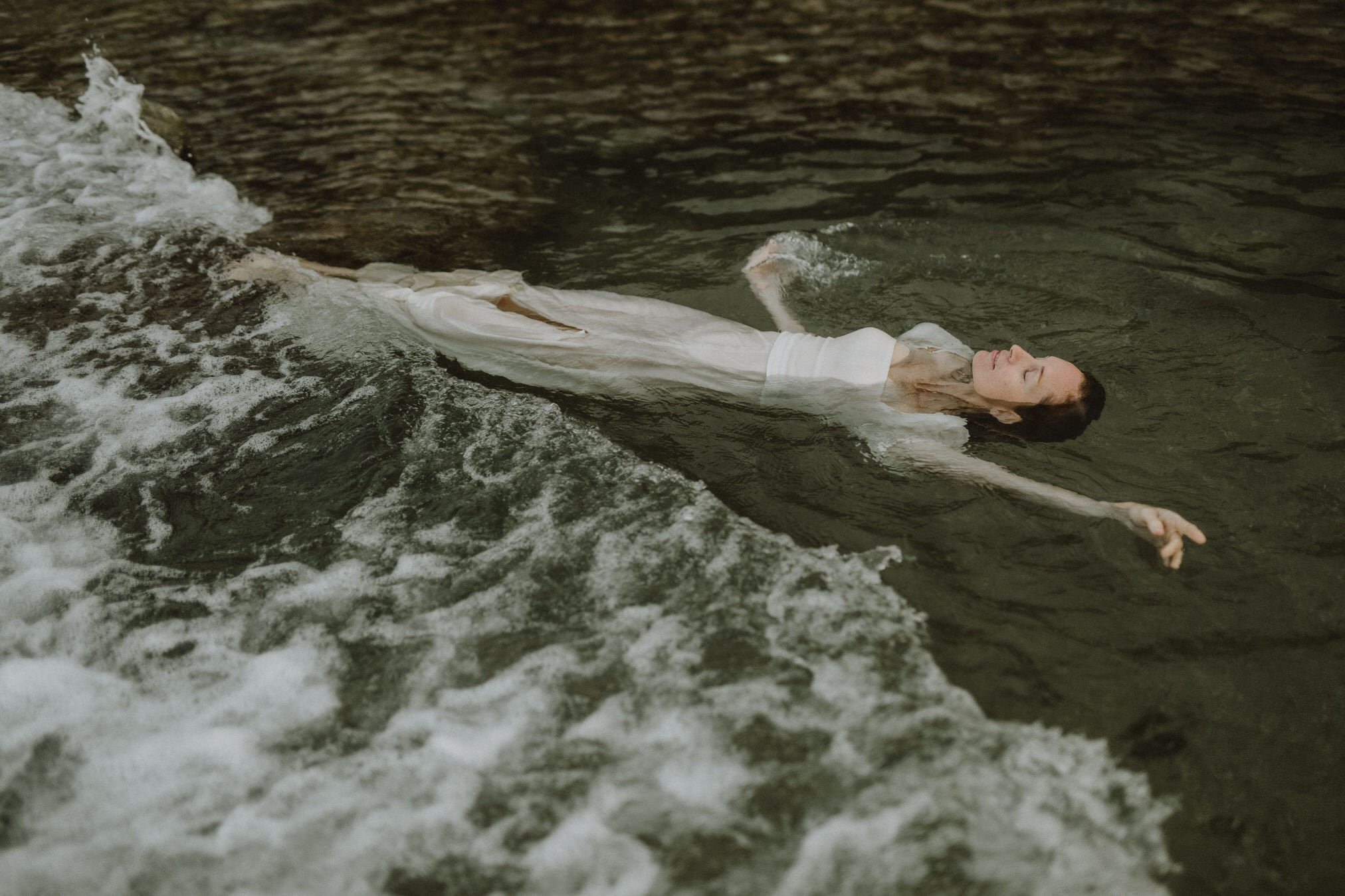 A woman in a long white dress floats on her back in dark moving water, her arms stretched wide as small waves break around her body. The soft light and muted colors give the scene a calm, suspended feeling, while the rushing water adds tension and movement around her still posture.