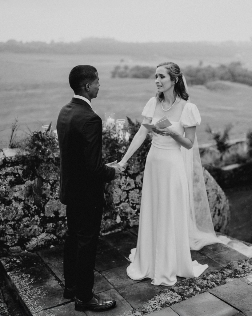 In a black-and-white photograph, a couple stands outdoors during a wedding ceremony, holding hands while the bride reads from a small notebook. The landscape behind them is soft and misty, and the quiet setting gives the moment an intimate, reflective feeling.