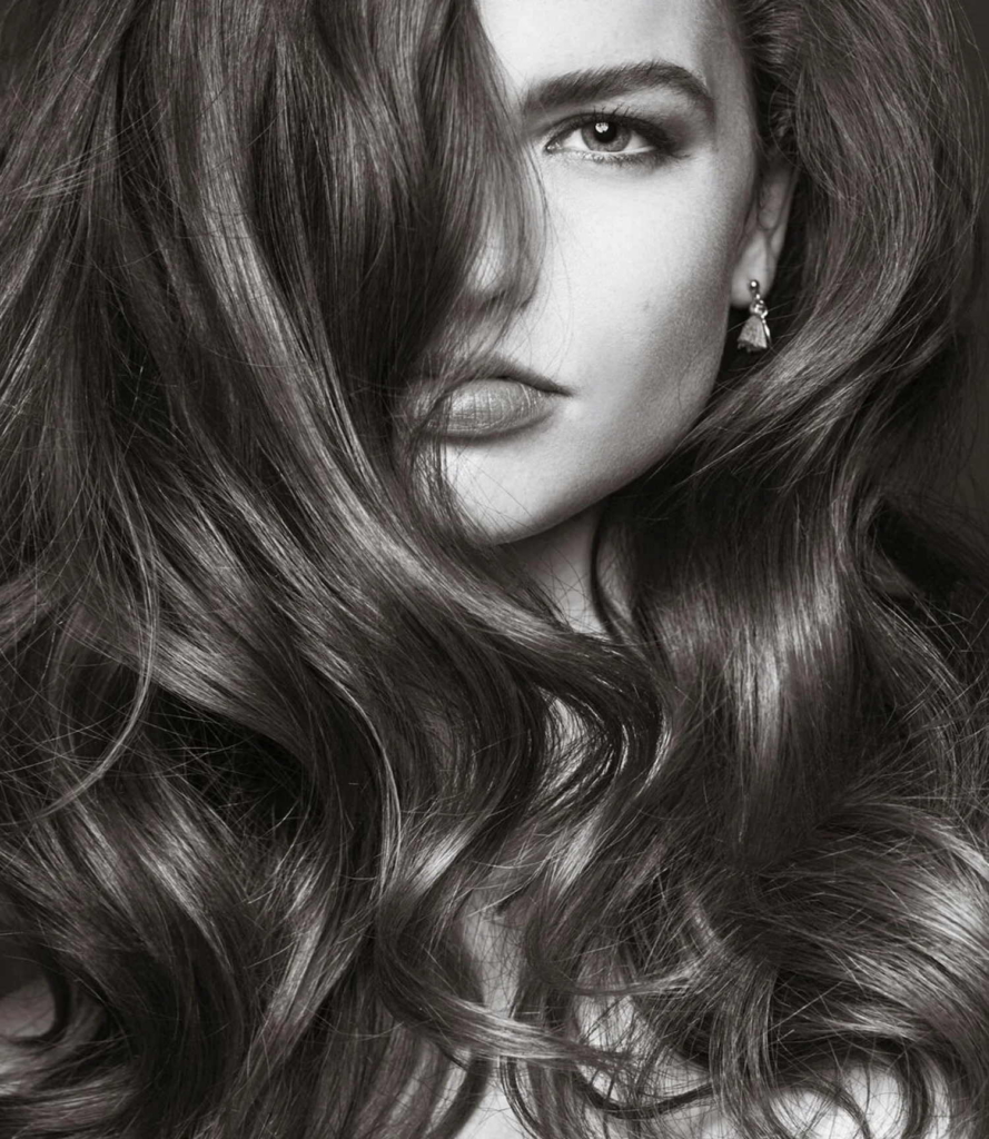 A black-and-white close-up portrait shows a woman with long wavy hair partially covering one eye.
Soft lighting emphasizes the texture and movement of her hair while her visible eye looks directly at the camera.