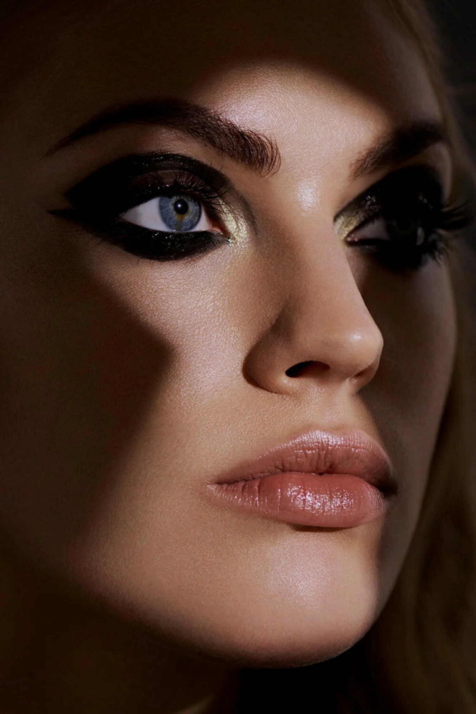 A close-up beauty portrait shows a woman with dramatic black eye makeup and glossy nude lips.
Soft lighting highlights the texture of her skin and the sharp contrast around her blue eyes.