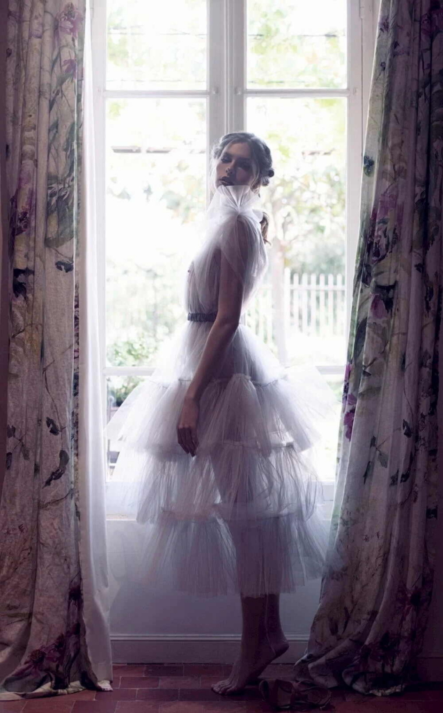 A woman in a layered tulle dress stands barefoot in front of a tall window framed by floral curtains.
Soft natural light filters through the room, creating a delicate and dreamlike fashion portrait.