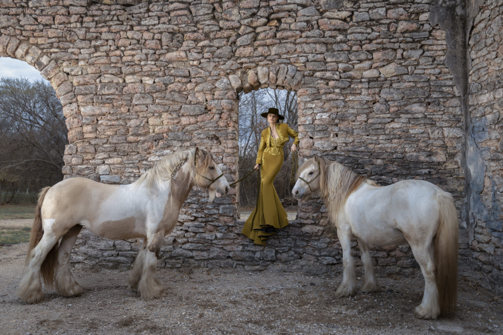 A series of fashion portraits show women posing with horses beside rustic stone ruins in warm natural light.
Flowing gowns, wide-brimmed hats, and textured landscapes create a cinematic Western-inspired atmosphere throughout the collection.