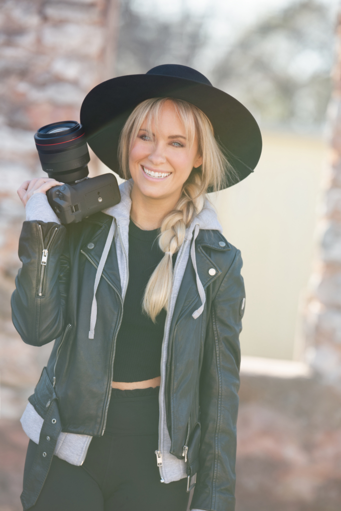 Photographer Dixie Dixon stands outdoors holding a professional camera in front of a rustic stone wall.
She wears a red leather jacket and cream beret, smiling down at the camera in soft natural light.