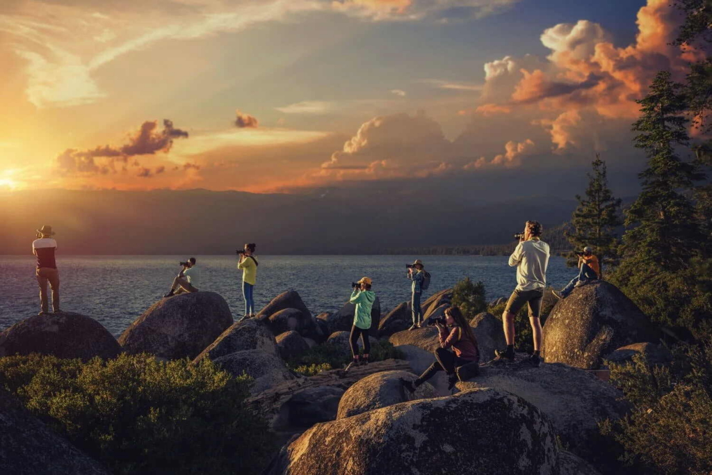 A group of photographers stands on large rocks beside a lake at sunset.
Dramatic orange and purple clouds fill the sky as each photographer points their camera toward the distant mountain landscape.