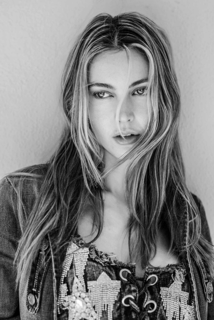 A black-and-white close-up portrait shows a young woman with long hair falling across part of her face.
She looks directly at the camera while soft lighting highlights her eyes and textured clothing.