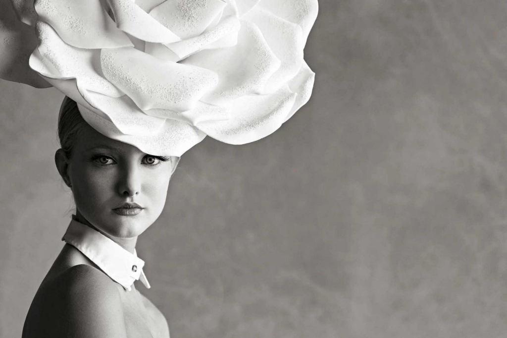A black-and-white portrait shows a woman wearing an oversized sculptural headpiece shaped like layered flower petals.
Soft studio lighting and a minimal background create an elegant and dramatic fashion composition.
