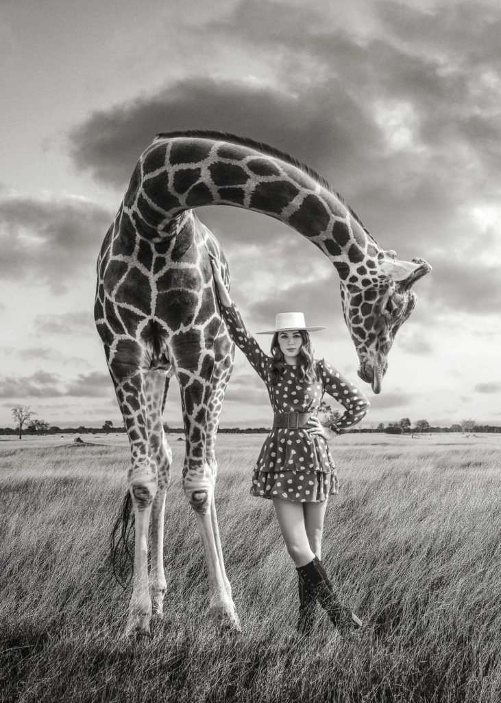 A black-and-white portrait shows a woman standing beside a giraffe in an open grassy landscape.
The giraffe bends its long neck gracefully over her as dramatic clouds fill the sky behind them.