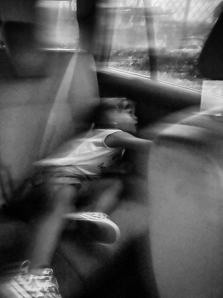Dreamlike black and white photograph of children resting inside a moving vehicle, layered with reflections and motion blur that create a fragmented, memory-like atmosphere.
