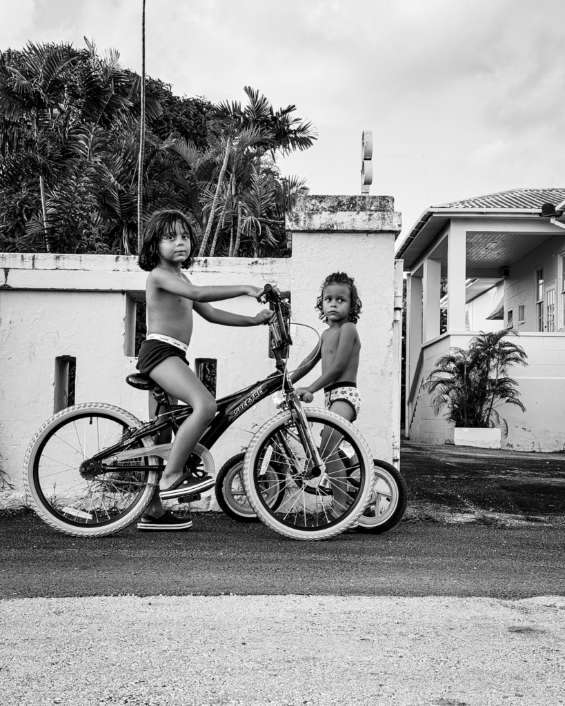 Two young children stand with their bicycles on a wet residential street beside a white wall and tropical plants. Both are shirtless and looking directly toward the camera, while the cloudy sky and damp pavement give the scene a calm, still atmosphere.