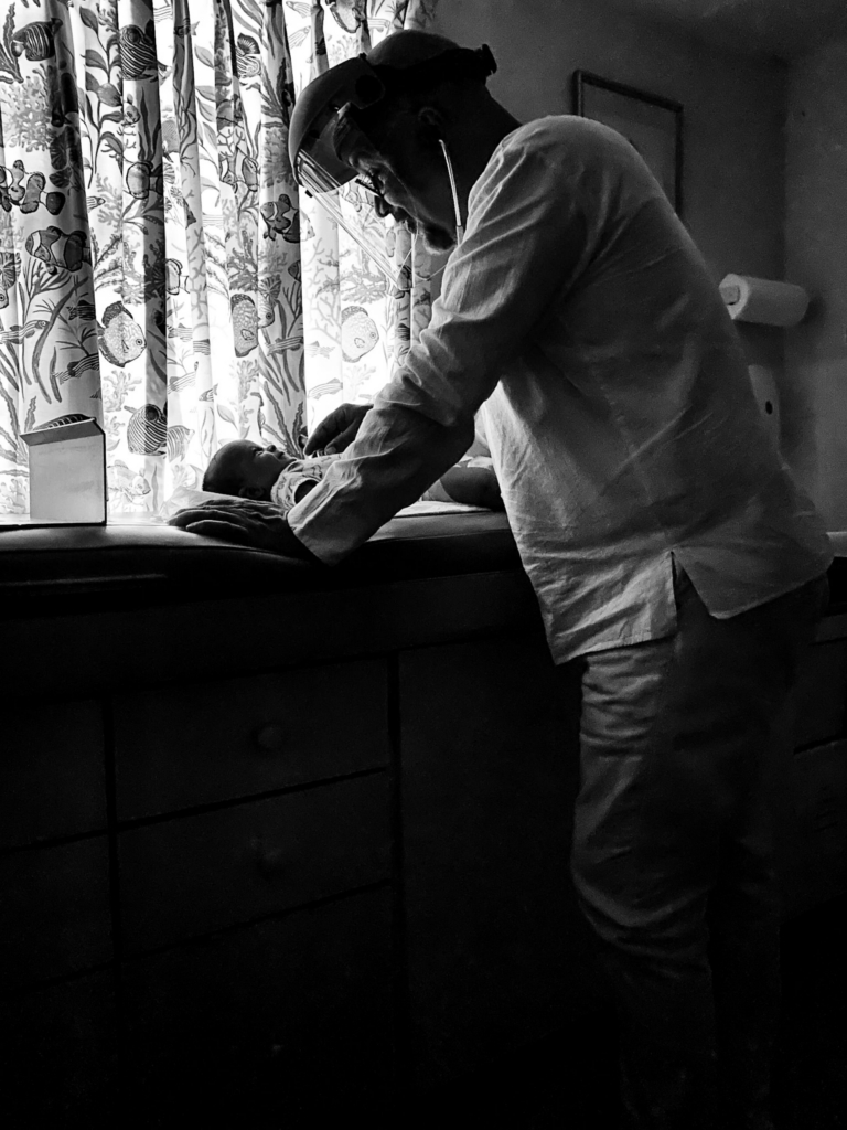A man wearing a face shield bends over a baby lying on a changing table near a softly lit window with patterned curtains. The black-and-white lighting and careful posture suggest a quiet moment of care and attention in a dim interior space.