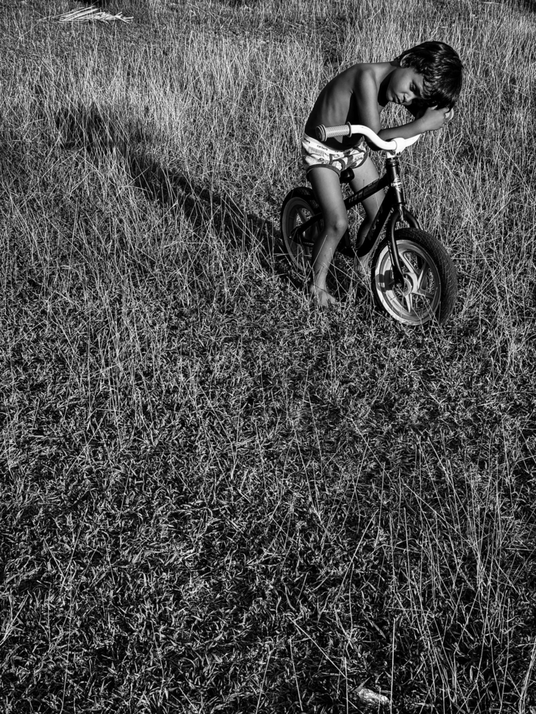 A young child stands barefoot in a dry grassy field beside a small balance bike, leaning their head against one arm on the handlebars. Strong sunlight creates deep shadows across the textured grass, and the child’s posture feels quiet and tired in the open landscape.