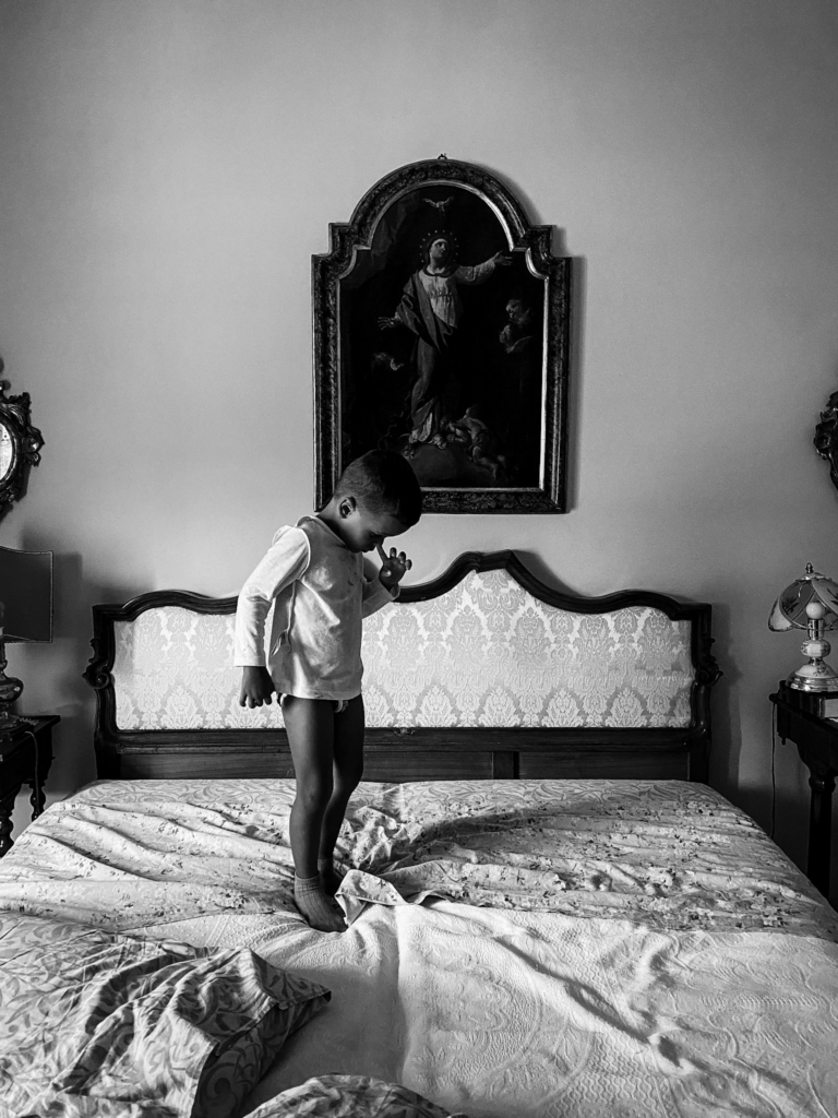 In a black-and-white image, a small child stands alone on a large bed in an old-fashioned room with patterned fabric and antique furniture. A framed religious painting hangs above the headboard, and the child’s downward gaze and still posture create a quiet, contemplative mood.