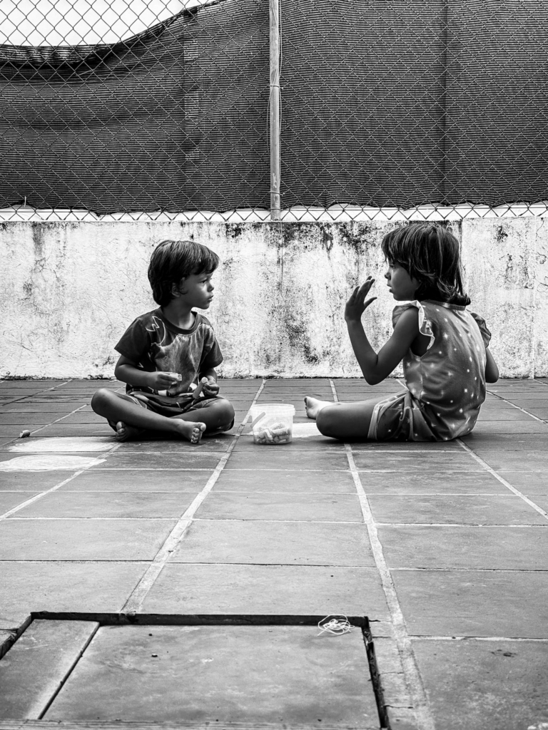 Black and white documentary photograph of two young children sitting on a tiled floor sharing a quiet moment together.