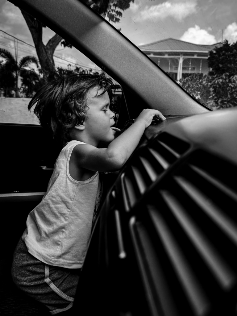 A young child leans forward inside a car, reaching toward the dashboard with one hand while looking out through the windshield. The black-and-white tones and close perspective emphasize the child’s concentration and curiosity inside the small enclosed space.