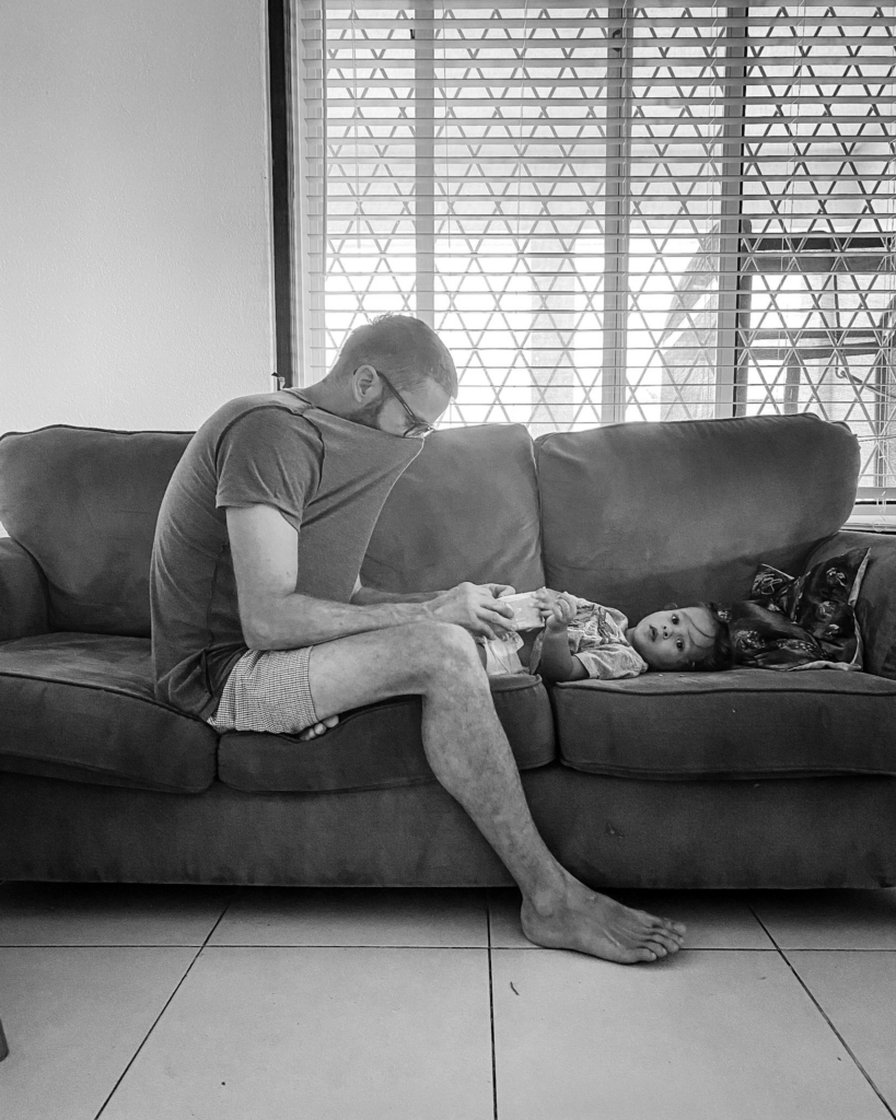 In a black-and-white photograph, a man sits barefoot on a couch feeding a young child lying beside him while soft daylight filters through the window behind them. The quiet posture and simple living room setting give the scene a gentle, everyday intimacy.