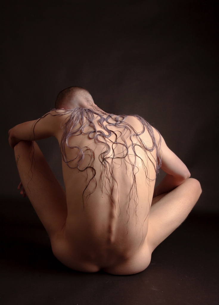 Conceptual nude portrait with organic hair-like textures and introspective sculptural posture.