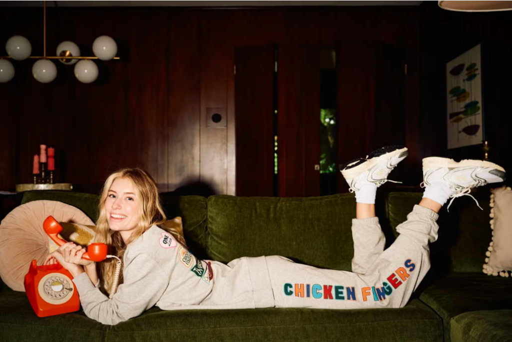 A smiling young woman lies on a green velvet couch while holding a bright orange rotary phone.
She wears a matching gray lounge set and sneakers, creating a playful retro-inspired scene with warm indoor lighting.