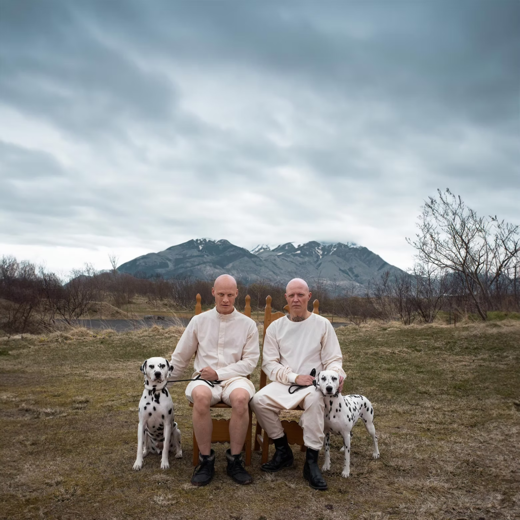 Two bald individuals sit side by side on wooden chairs in an open landscape, each holding a Dalmatian on a leash. Dressed in matching light-colored clothing, they face the camera with serious expressions, while mountains and a dramatic cloudy sky form a striking backdrop behind them.