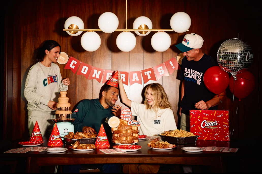 Four friends gather around a table filled with fried chicken, fries, and party decorations at a Raising Cane’s celebration.
Red balloons, party hats, and a “Congrats!” banner create a cheerful and festive atmosphere indoors.