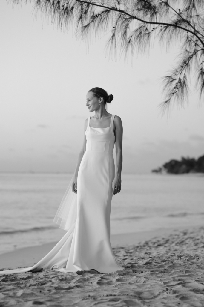 Bride in a minimalist wedding dress standing barefoot on a quiet beach at sunset.