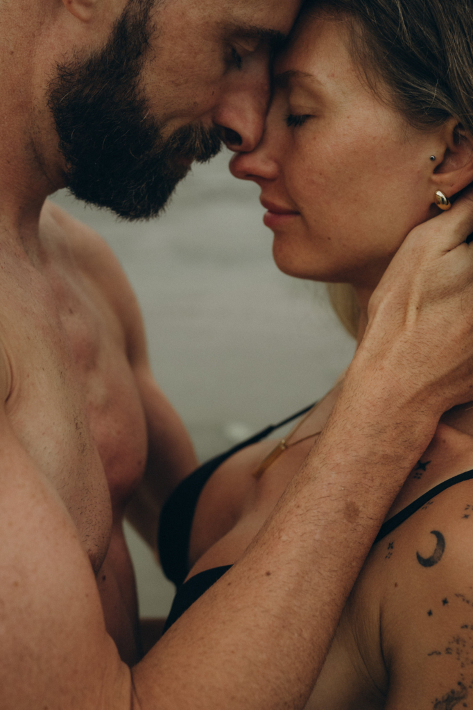 A shirtless man and a woman in a black swimsuit hold each other closely with their foreheads pressed together near the water. Their eyes are closed, and the warm muted light gives the scene a calm, tender feeling while small details like tattoos and jewelry remain visible.