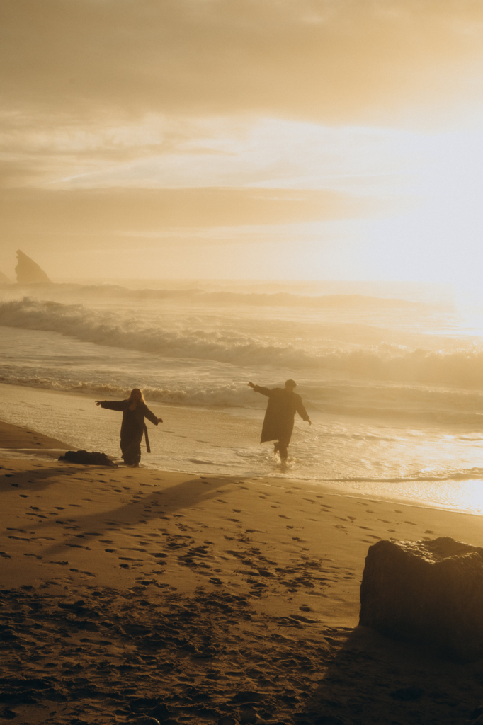 Two figures run and play at the edge of the ocean during golden sunset light, their dark silhouettes partially reflected on the wet sand. Waves roll in around them while long shadows and sea mist soften the beach into a warm, dreamlike scene.
