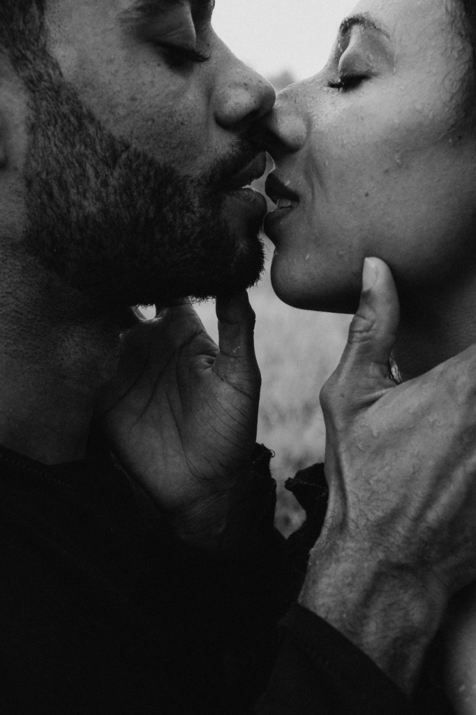 Two people are shown in an intimate close-up, their faces nearly touching as they kiss with their eyes closed. Water droplets and soft black-and-white tones emphasize the texture of their skin and hands, creating a quiet, deeply personal moment.