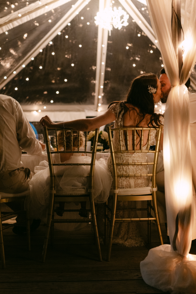 A bride and groom sit close together inside a softly lit reception tent, kissing beside a small child seated between them. Warm string lights and draped fabric surround the table, creating an intimate and quiet atmosphere during the celebration.