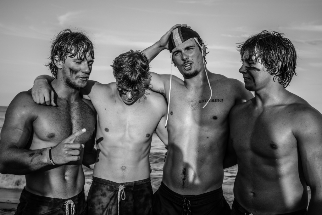 Four surf lifesaving athletes stand together on the beach after a race, their wet hair and sand-marked faces showing the effects of competition. The black-and-white portrait captures the group with arms around one another, with the ocean visible behind them in soft evening light.