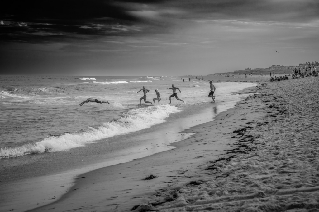A group of beach athletes runs into the ocean during a surf lifesaving event, captured along a wide stretch of shoreline. The black-and-white image emphasizes the dramatic sky, rolling waves, and the athletes' movement as they enter the water, with beachgoers visible in the distance.