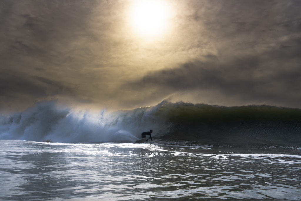 A surfer rides inside a breaking wave as sunlight filters through clouds above the ocean. The low-angle perspective from the water emphasizes the scale of the wave, with spray and reflections shimmering across the surface.