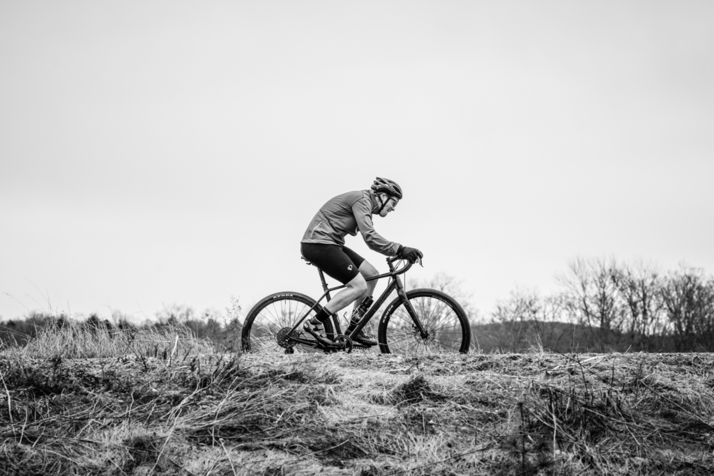A cyclist rides across a gravel trail in an open landscape, captured in profile against a wide, empty sky. The black-and-white image emphasizes the rider’s posture and the textured terrain, with bare trees lining the distant horizon.