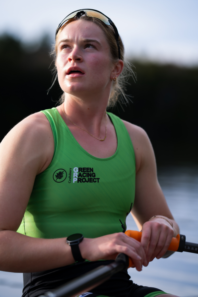 A female rowing athlete from the Green Racing Project sits in a racing shell, holding an oar while looking upward. Soft natural light illuminates her face and green racing uniform, with calm water and a blurred shoreline in the background.