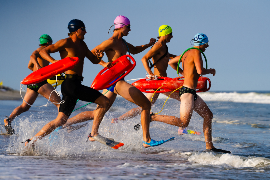 Lifeguard competition athletes sprint from the shoreline into the ocean carrying bright red rescue buoys. The competitors wear swim caps and fins as water splashes around their legs, with waves and a clear blue sky forming the background.