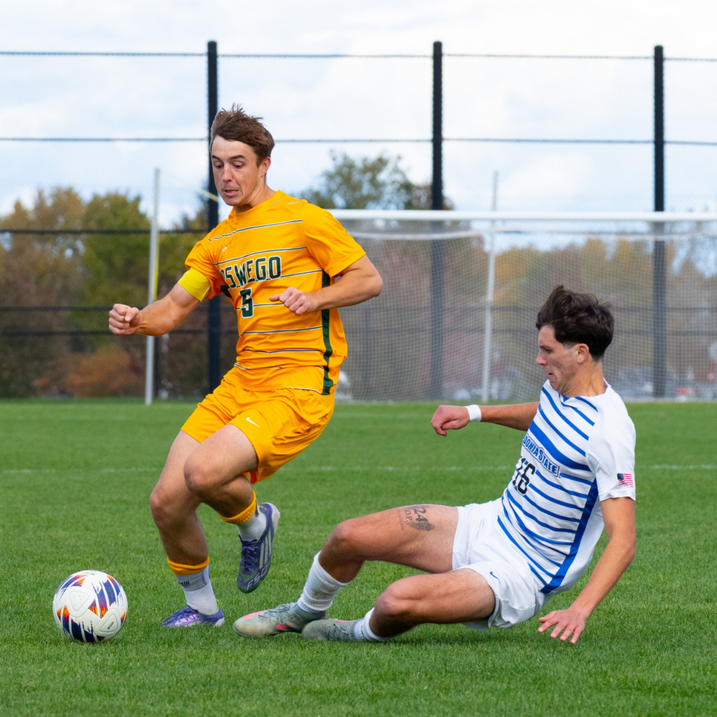 Two soccer players compete for possession of the ball during a college match, with one player challenging while the other attempts to maintain control. The action takes place on a grass field with the goal and surrounding landscape softly visible in the background.