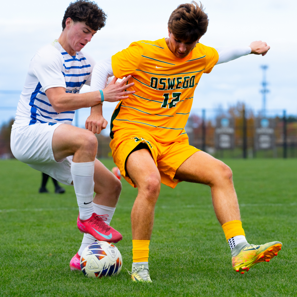 Two soccer players compete for possession of the ball during a college match, with one player challenging while the other attempts to maintain control. The action takes place on a grass field with the goal and surrounding landscape softly visible in the background.