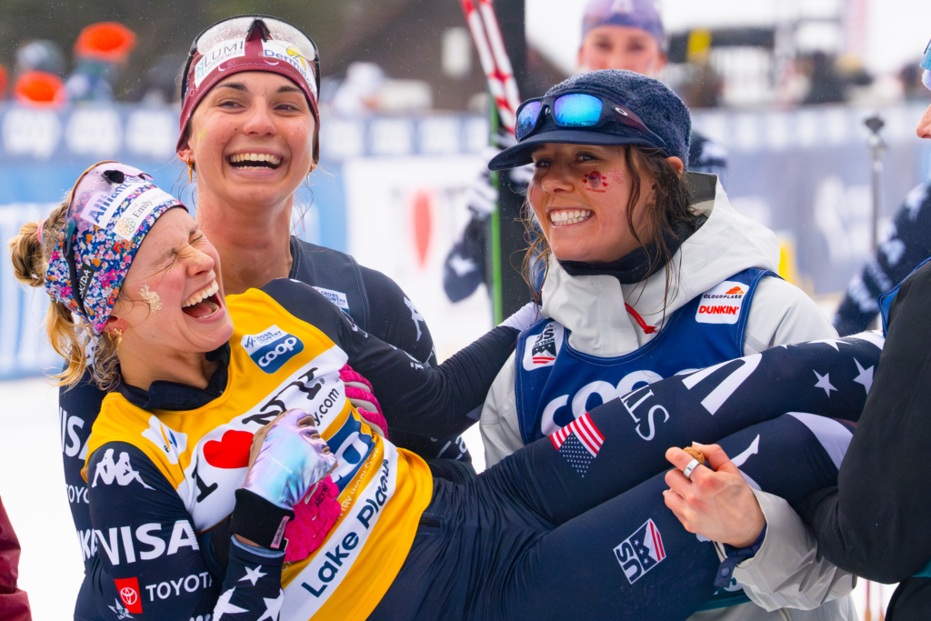 Three cross-country skiing athletes celebrate together after a race, smiling and laughing in a snowy competition venue. Wearing race bibs, winter gear, and team uniforms, they stand closely together while one athlete is lifted in the arms of her teammates.