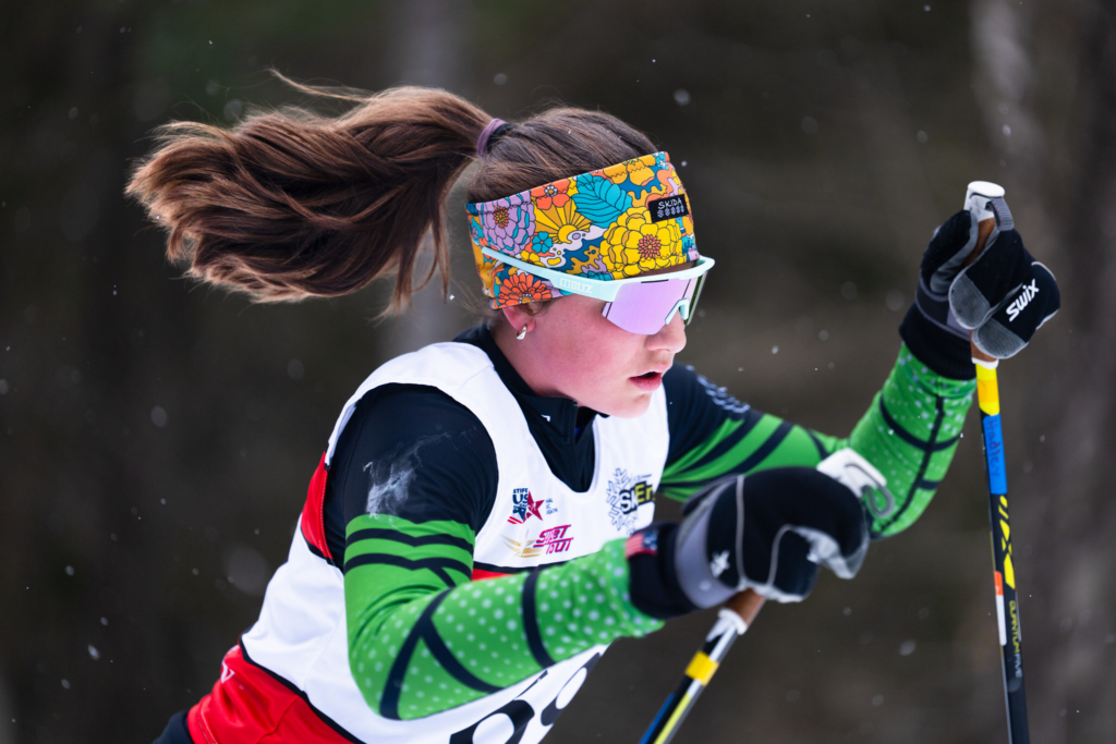 A female cross-country skier climbs a snowy section of the trail during a race, driving her poles into the snow with focused effort. She wears a patterned headband, reflective sunglasses, and a race bib, while light snow falls in front of a dark woodland backdrop.