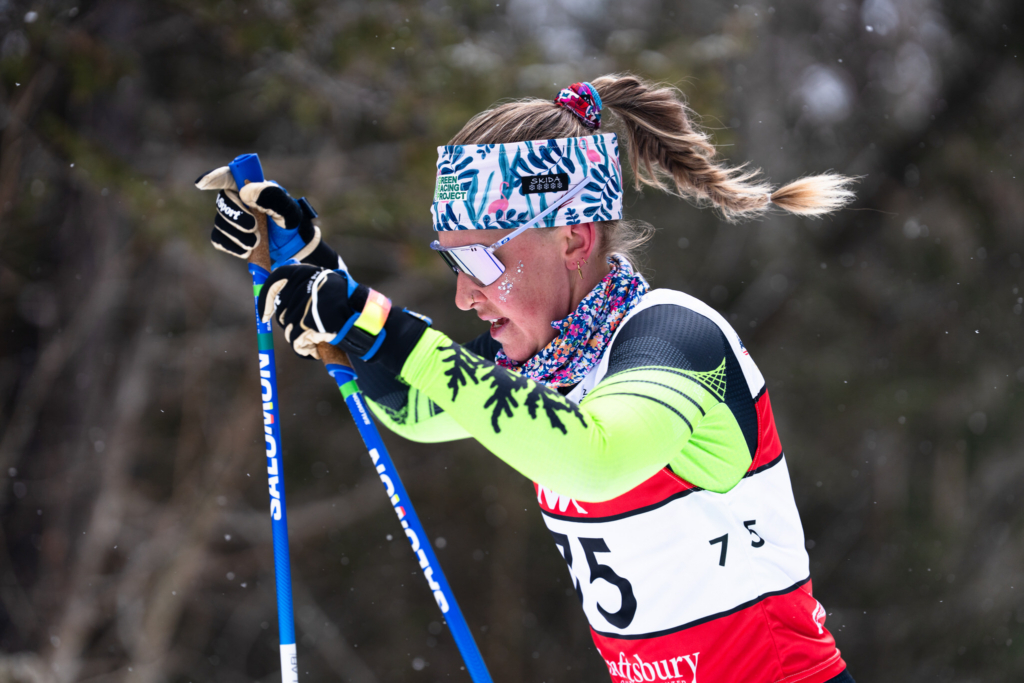 A cross-country skier races through a snowy course, pushing forward with ski poles as her ponytail swings behind her. She wears mirrored sunglasses, a colorful headband, and a race bib, with soft snowflakes visible against a blurred forest background.