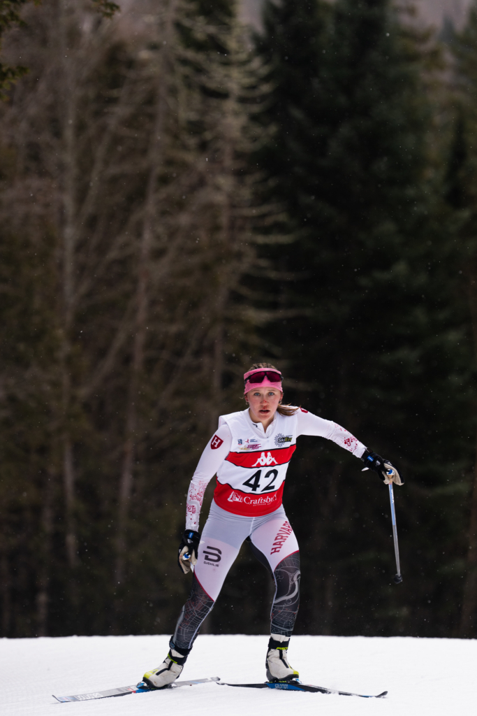 A female cross-country skier glides across a snow-covered trail during a race, wearing a numbered bib and pink headband. Light snowflakes drift through the air as a dark forest backdrop isolates the athlete against the bright snow.