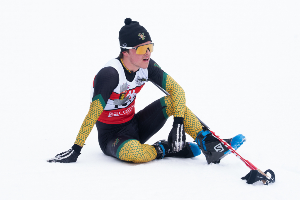 A cross-country skier sits on the snow after a race, adjusting a ski boot while holding a ski pole beside him. Wearing a race bib, winter hat, and reflective sunglasses, he is isolated against a bright, snow-covered background.