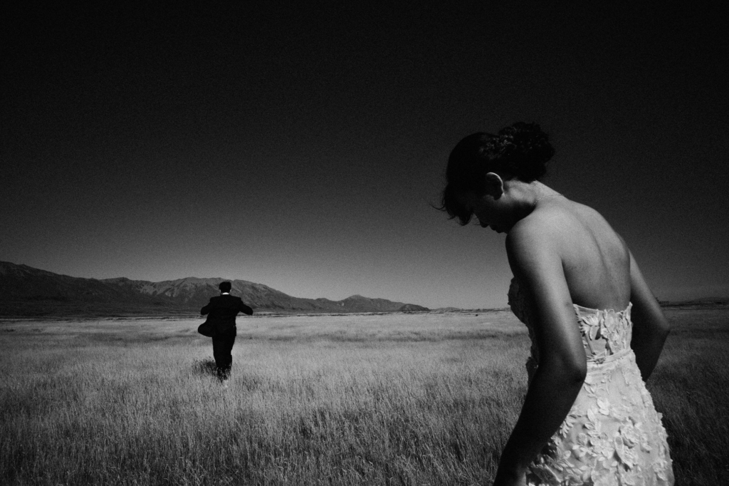 A black-and-white photograph shows a woman in a textured wedding dress standing in the foreground of a wide grassy field, her head tilted downward as she looks toward the ground. In the distance, another person in dark clothing walks away across the landscape, with mountains stretching along the horizon beneath a clear sky, creating a quiet and expansive atmosphere.