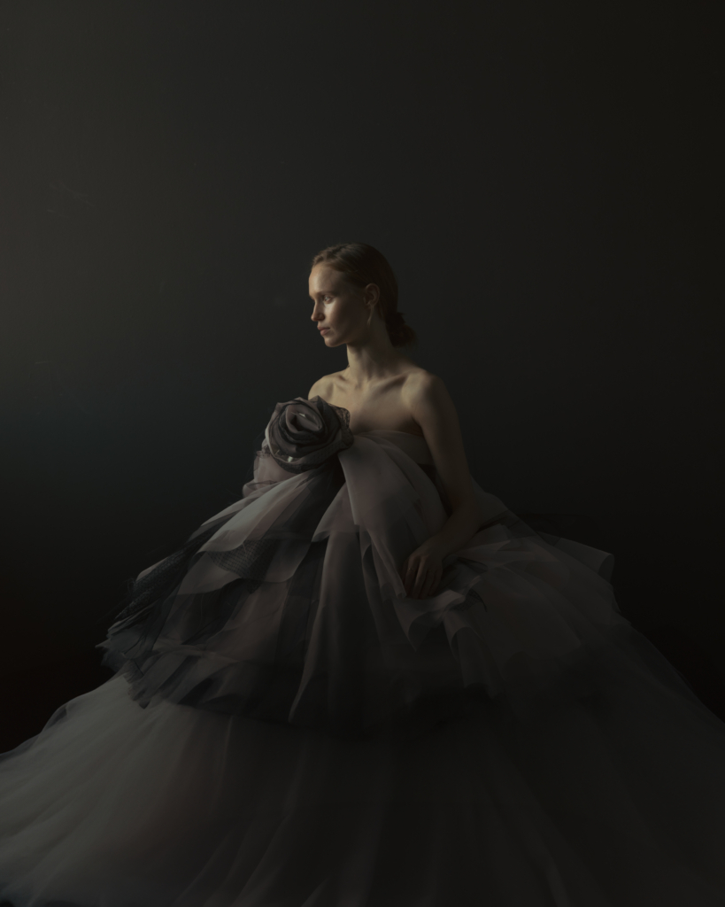 A woman sits in a dramatic sculptural gown made of layered translucent fabric, softly illuminated against a dark background. Looking away from the camera, she appears calm and reflective, while the gentle light and flowing textures create an elegant and intimate atmosphere.
