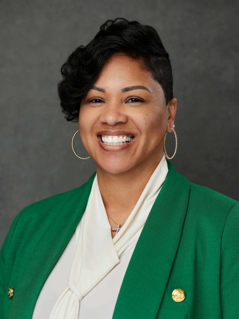 A woman smiles directly at the camera in a studio headshot with a gray background. She wears a green blazer over a white blouse, large hoop earrings, and a necklace, with soft lighting highlighting her face.